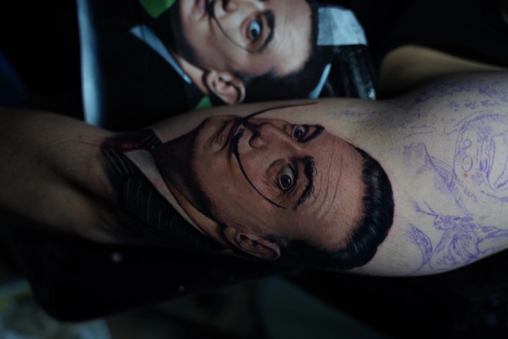 Colour portrait of Salvador Dalí tattooed on an upper arm, held next to the printed reference photo.
