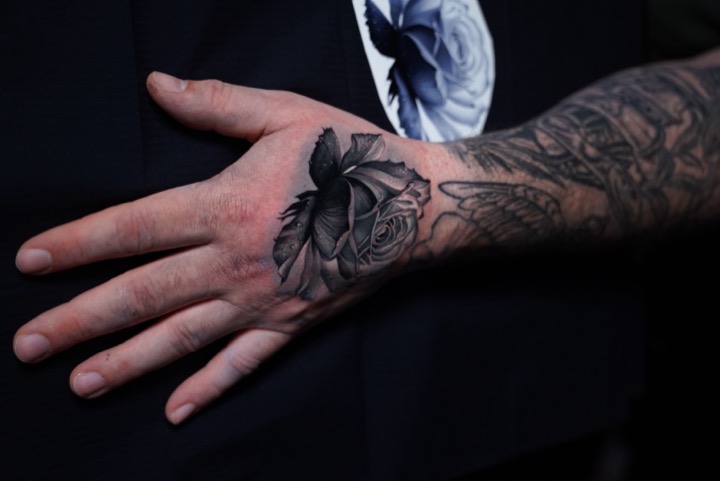 Black and grey rose tattooed on the back of a hand, the drawn design visible beside it.