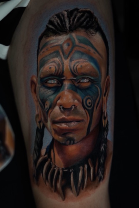 Colour portrait of a man with blue face paint, braids and a bone necklace, on an upper arm.